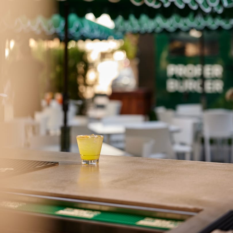 A yellow cocktail with ice sits on a bar, with the PROPER BURGER sign in the background.