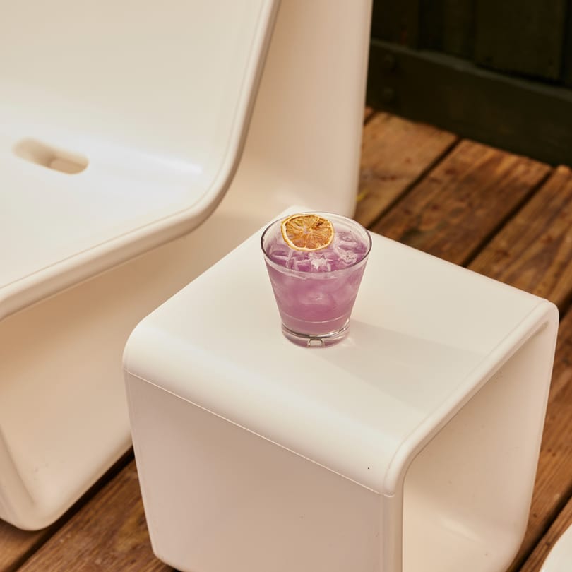 Purple cocktail with ice and a dried orange slice on a white table, with wooden floor.