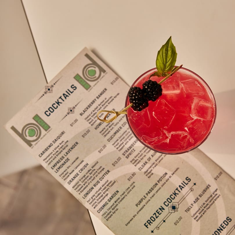 A vibrant red cocktail with blackberries and a basil leaf garnish sits next to a cocktail menu.