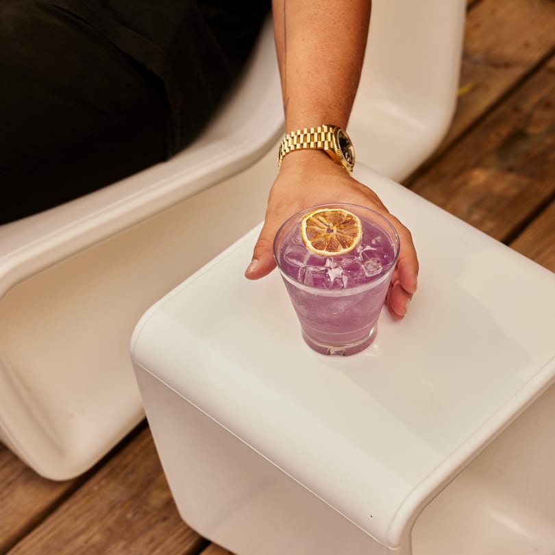 A hand with a gold watch reaches for a purple cocktail garnished with a dried orange slice.