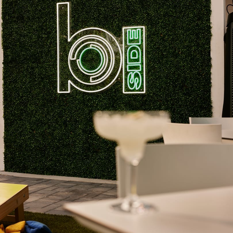 Neon "b-side" sign on a green hedge wall with a blurred cocktail glass in the foreground.