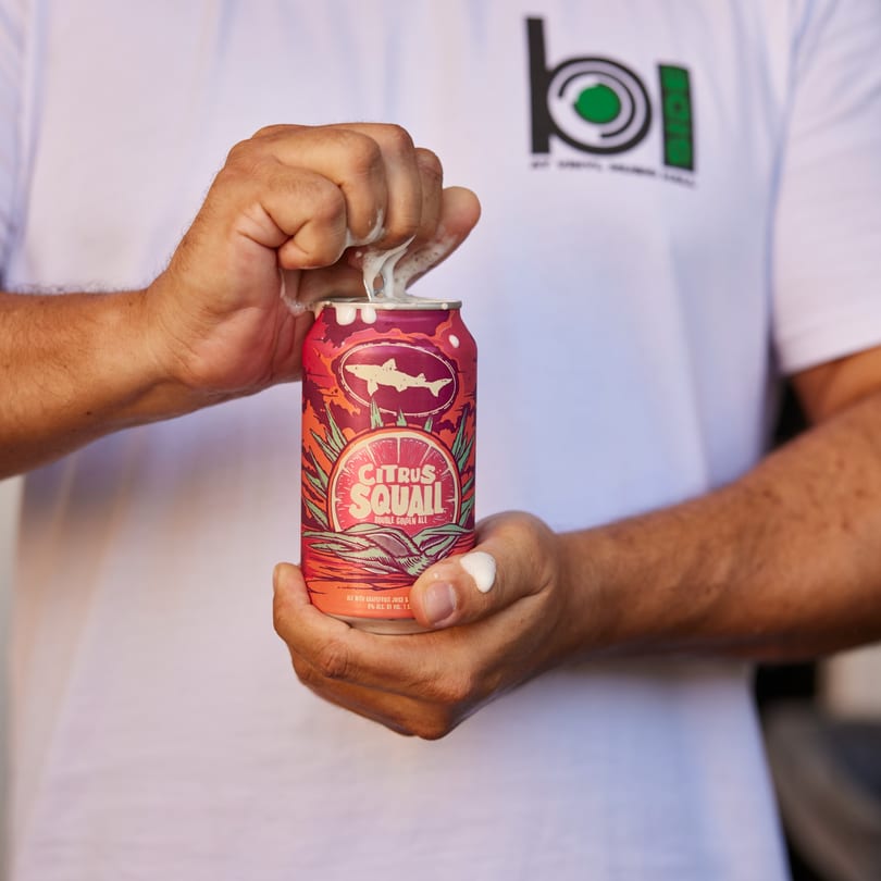 Hands holding a red "Citrus Squall Double Golden Ale" can, overflowing with foam.