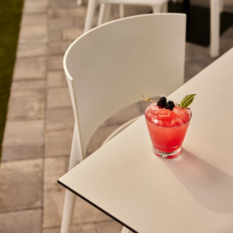 A vibrant red cocktail with blackberries and a basil leaf sits on a white outdoor table.