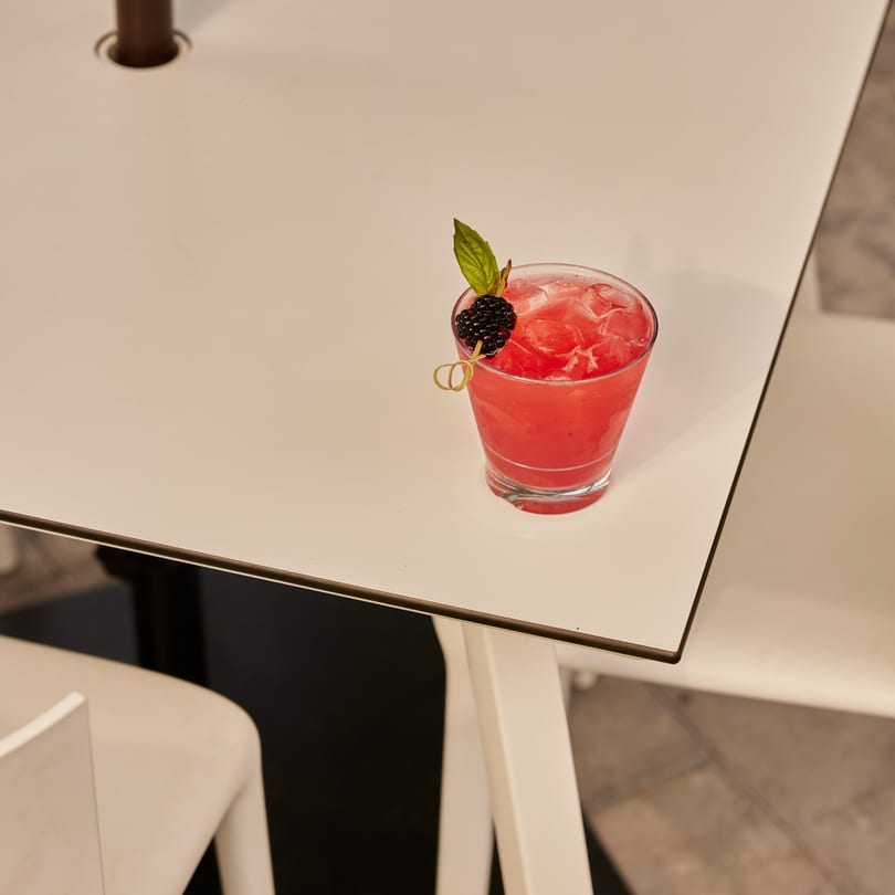 A vibrant red cocktail with ice, garnished with a blackberry and a green leaf, on a white table.