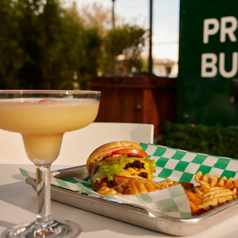 A PROPER BURGER with waffle fries and a cocktail on a white table outdoors.
