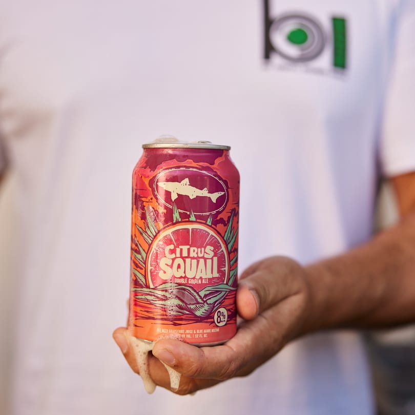 A hand holds a vibrant red and orange can of Citrus Squall Double Golden Ale, with foam spilling.