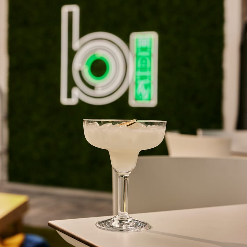 A margarita with a lime wedge on a white table, with a green neon sign in the background.