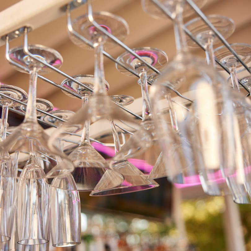Upturned wine and cocktail glasses hang from a rack above a bar with pink lighting.