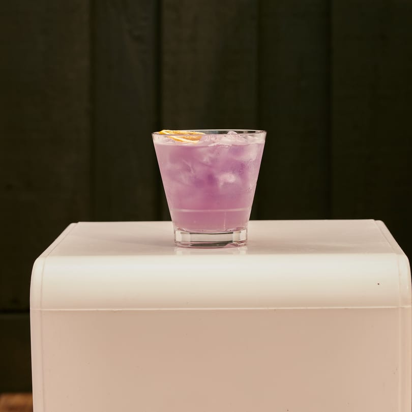 A vibrant purple cocktail with ice and an orange slice, on a white surface.