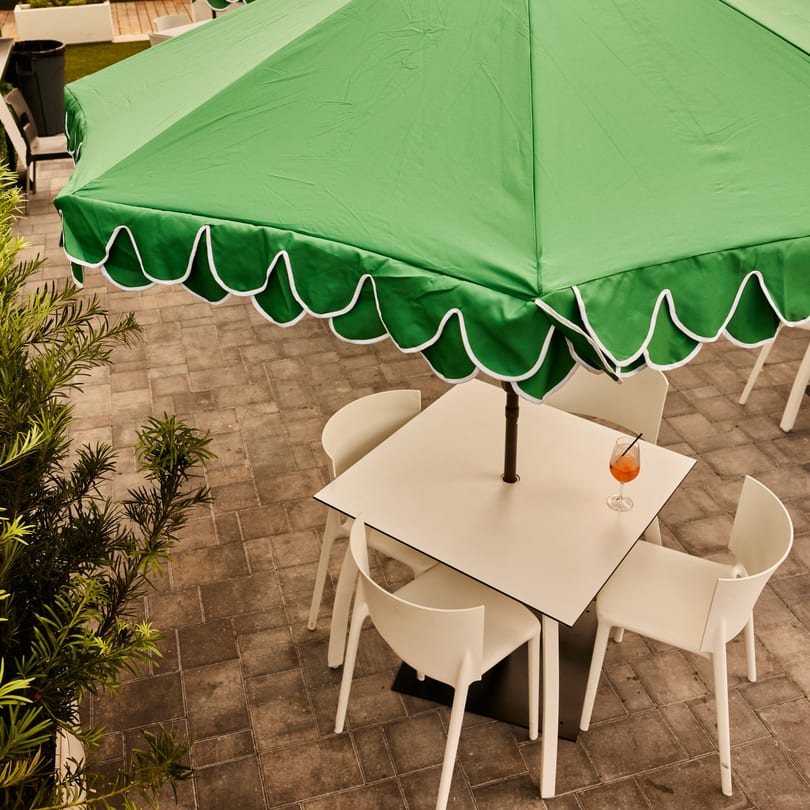 Overhead view of an outdoor dining table with white chairs, a green umbrella, and an orange drink.