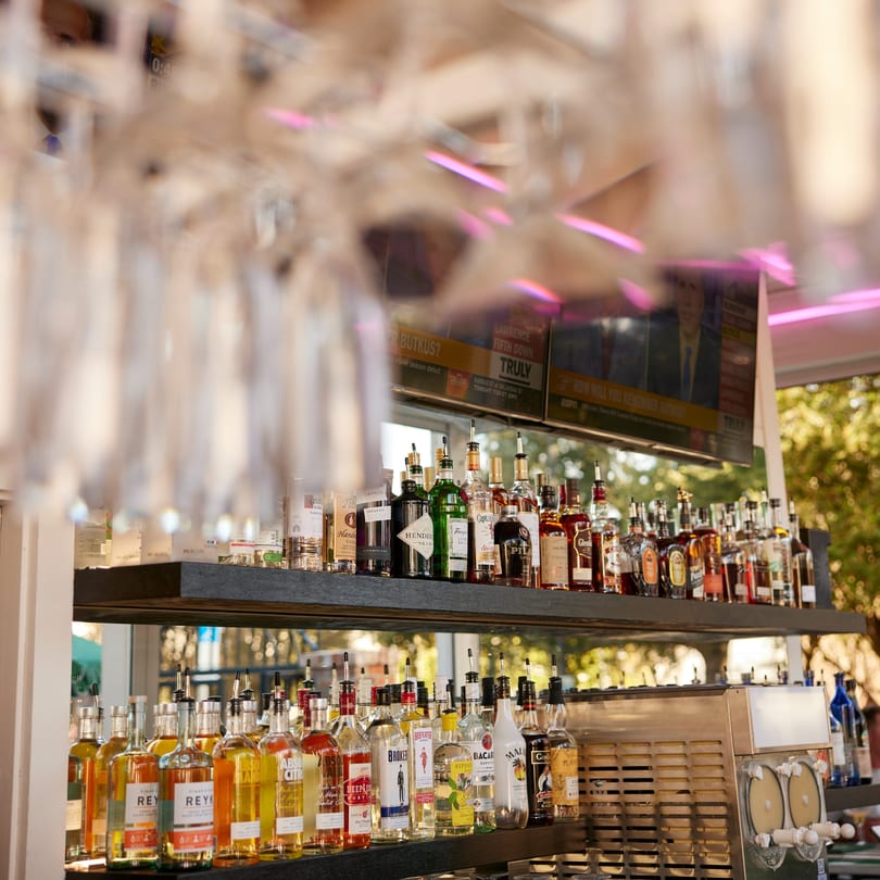 A bar at PROPER BURGER Downtown with liquor bottles, hanging glasses, and a TV screen.