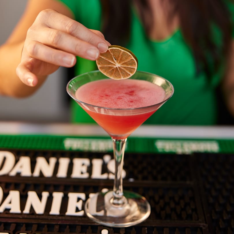 A hand garnishes a pink cocktail in a martini glass with a dried citrus slice.