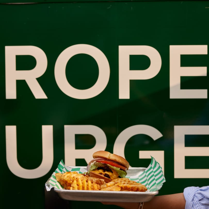 A Proper Burger with waffle fries on a tray, held in front of a green sign.