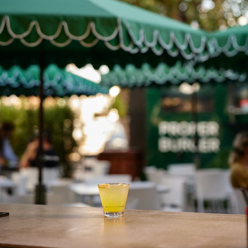 A yellow cocktail on a bar counter with green umbrellas and the "PROPER BURGER" sign in the background.