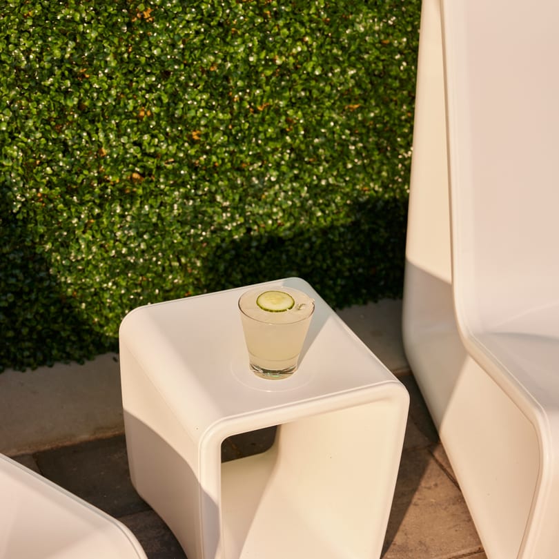 A refreshing cocktail with a cucumber slice sits on a white outdoor table next to a green hedge.