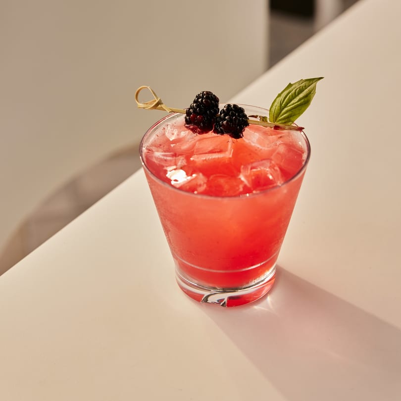 A vibrant red cocktail with ice, garnished with two blackberries and a basil leaf on a white surface.