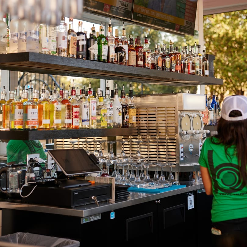 A bartender works at an outdoor bar with shelves of liquor bottles and a frozen drink machine.