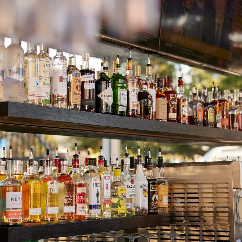 Two shelves of various liquor bottles with pour spouts, with a TV screen visible above.